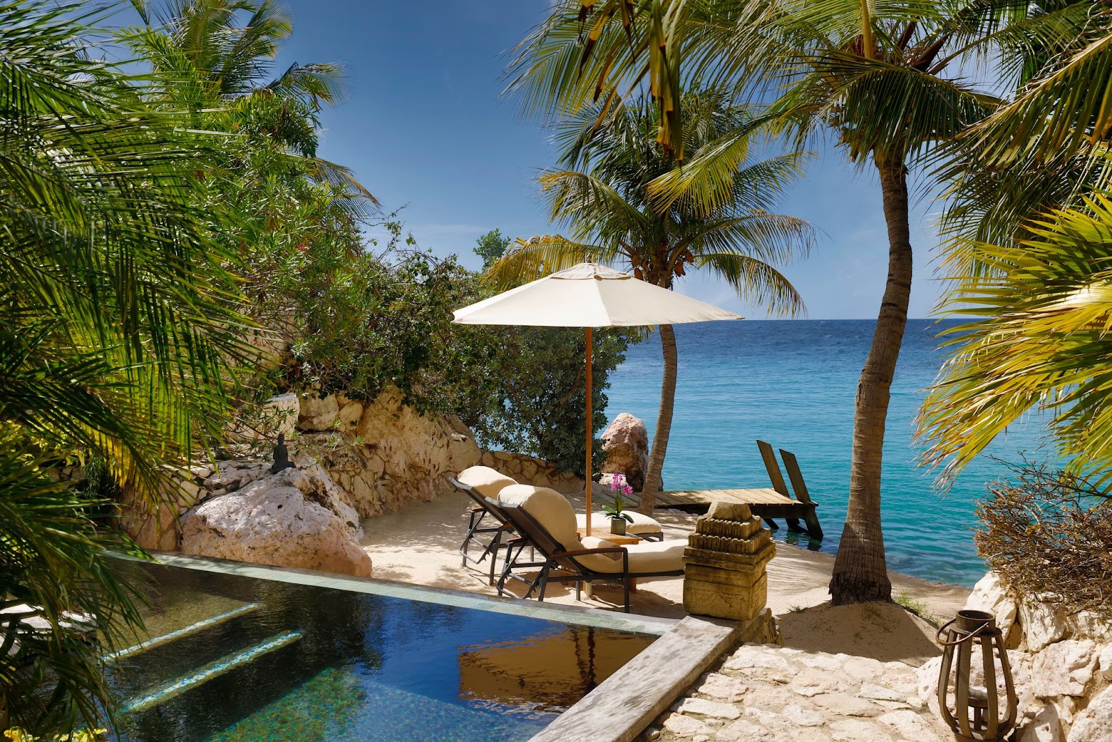 Beachfront lounge area at Baoase Luxury Resort with sunbeds under a white umbrella, rocky outcrops, palm trees, and turquoise Caribbean Sea.