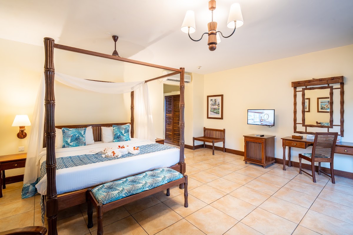 Spacious resort bedroom with four-poster wooden bed, blue patterned pillows, cream walls, tile flooring, work desk with TV, and wooden furniture throughout.
