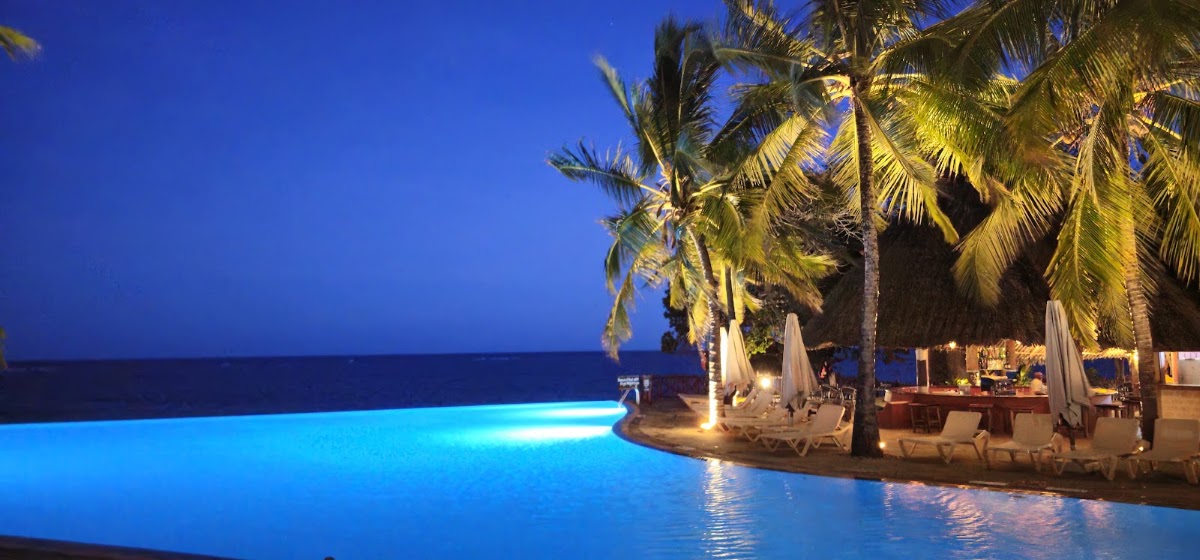 Illuminated infinity pool overlooking the ocean at dusk, with palm trees and lounge seating along the deck at Baobab Beach Resort & Spa.