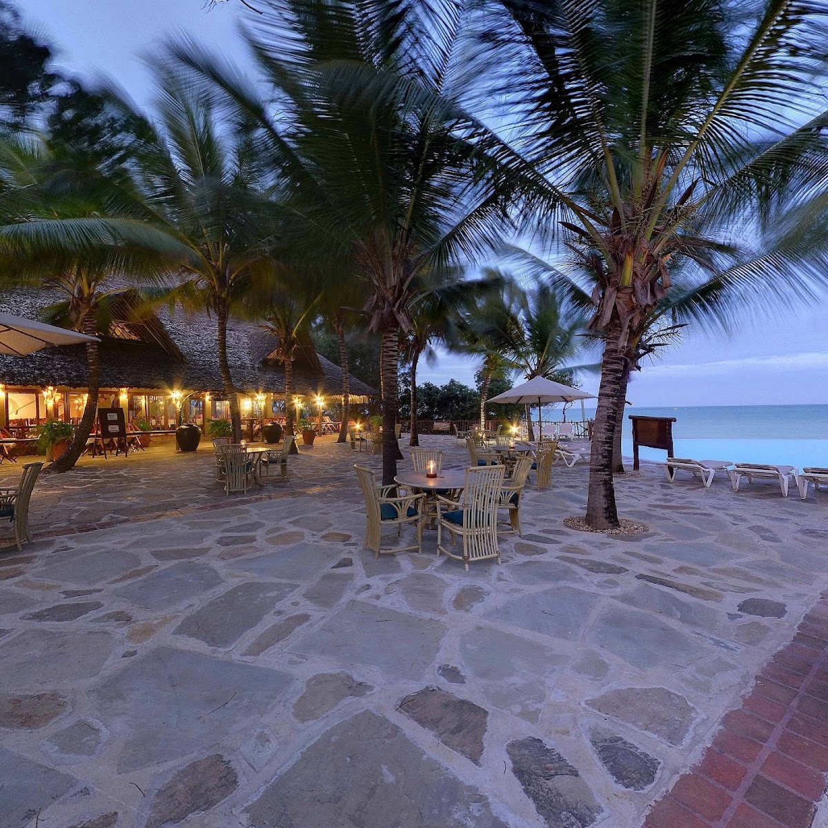 Beach terrace at dusk with lit dining tables among palm trees, illuminated lodge building, and ocean view beyond.