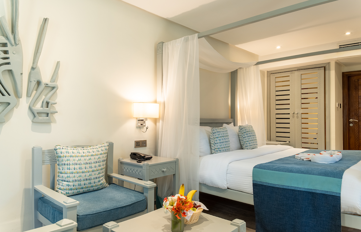 Modern resort bedroom with king bed, sheer canopy curtains, blue and white upholstered seating area, and fresh flowers on side table.