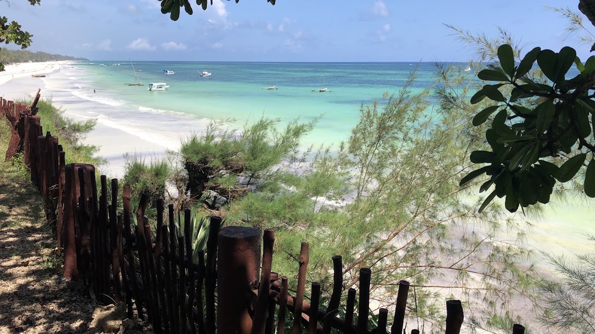White sand beach with turquoise water and moored boats at Baobab Beach Resort & Spa, framed by tropical vegetation and weathered wooden pilings.