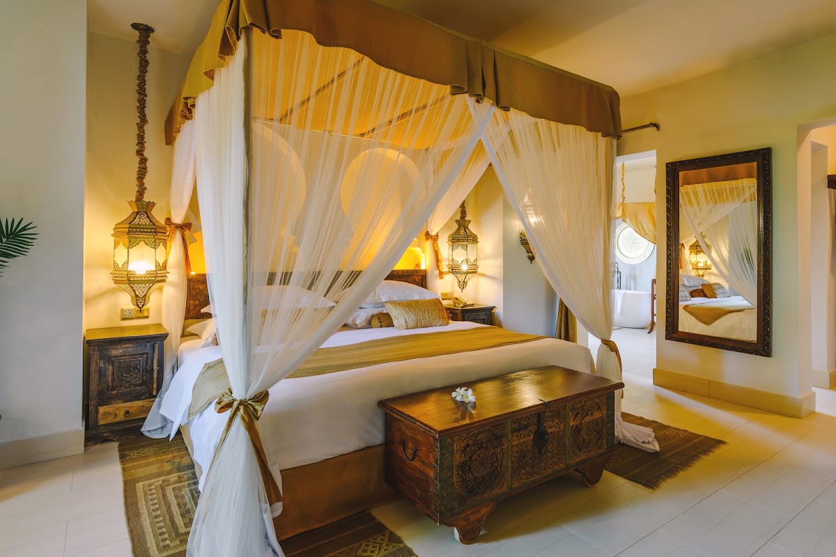 Resort bedroom with a four-poster canopy bed draped in white curtains, ornate carved wooden chest, brass lanterns, and Moroccan-style décor in warm gold and cream tones.