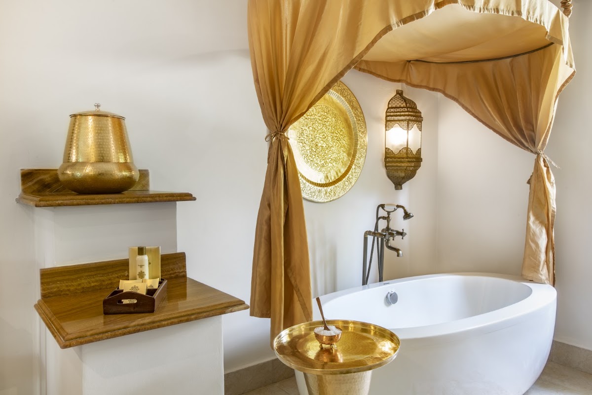 Luxury bathroom with freestanding white tub, gold draped canopy, brass accessories, and ornamental wall decor at Baraza Resort & Spa.