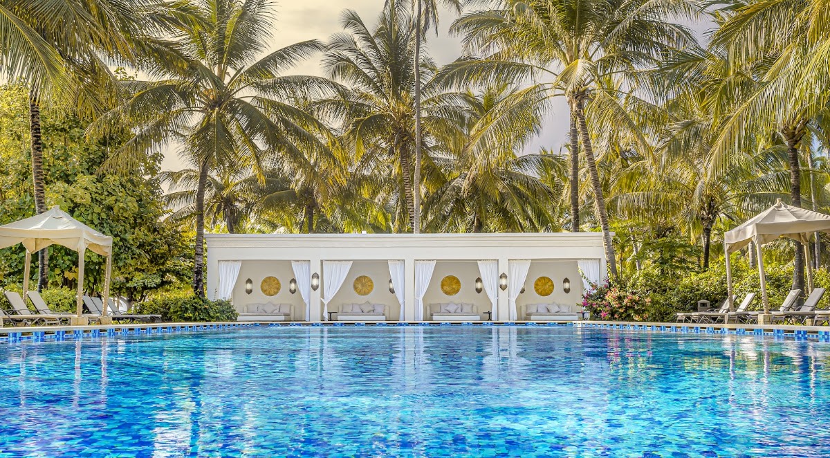 Resort swimming pool with white cabanas and gold circular accents, surrounded by palm trees and lounge chairs with umbrellas.