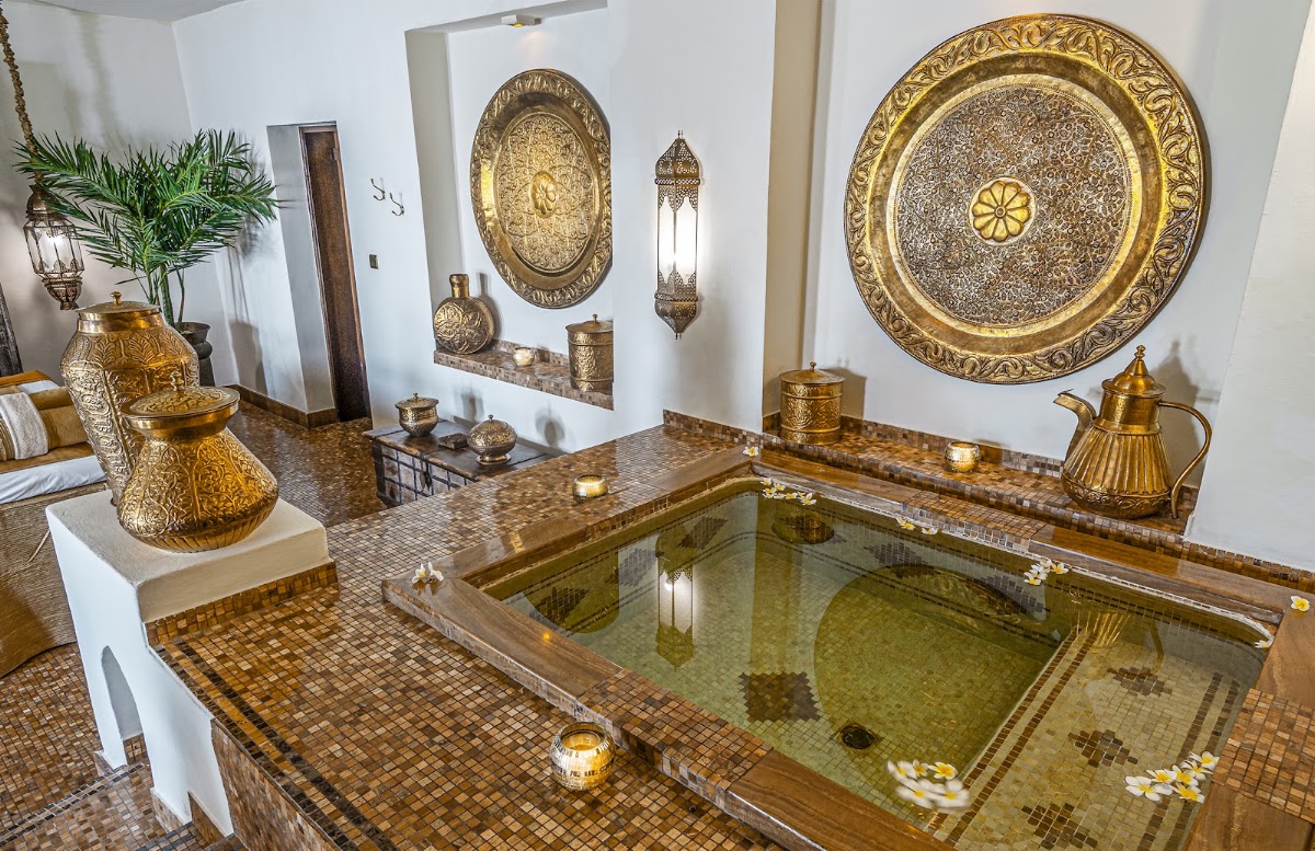 Sunken bath with green tiled interior surrounded by ornate brass vessels and circular wall plates in Moroccan-style spa room.