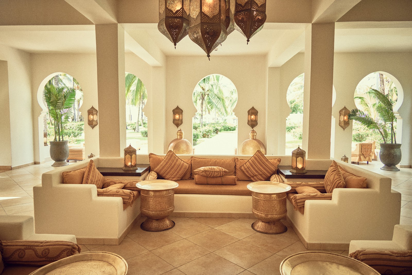 Cream-colored lounge with arched windows framing palm trees, tan seating, brass lanterns, and geometric pendant lights overhead.