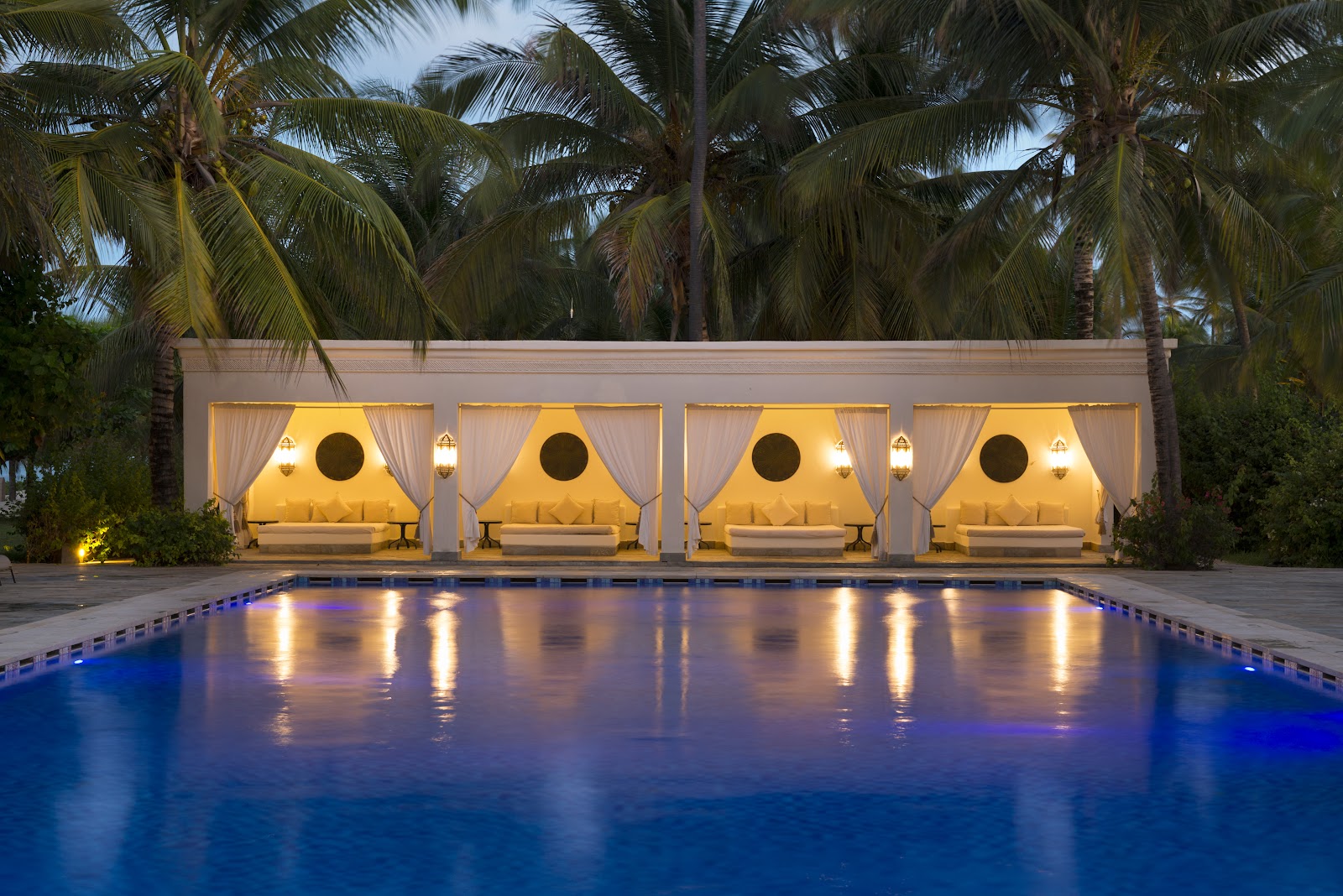 White Moroccan-style courtyard with arched colonnades, brass lanterns, woven poufs, and palm trees under clear sky.