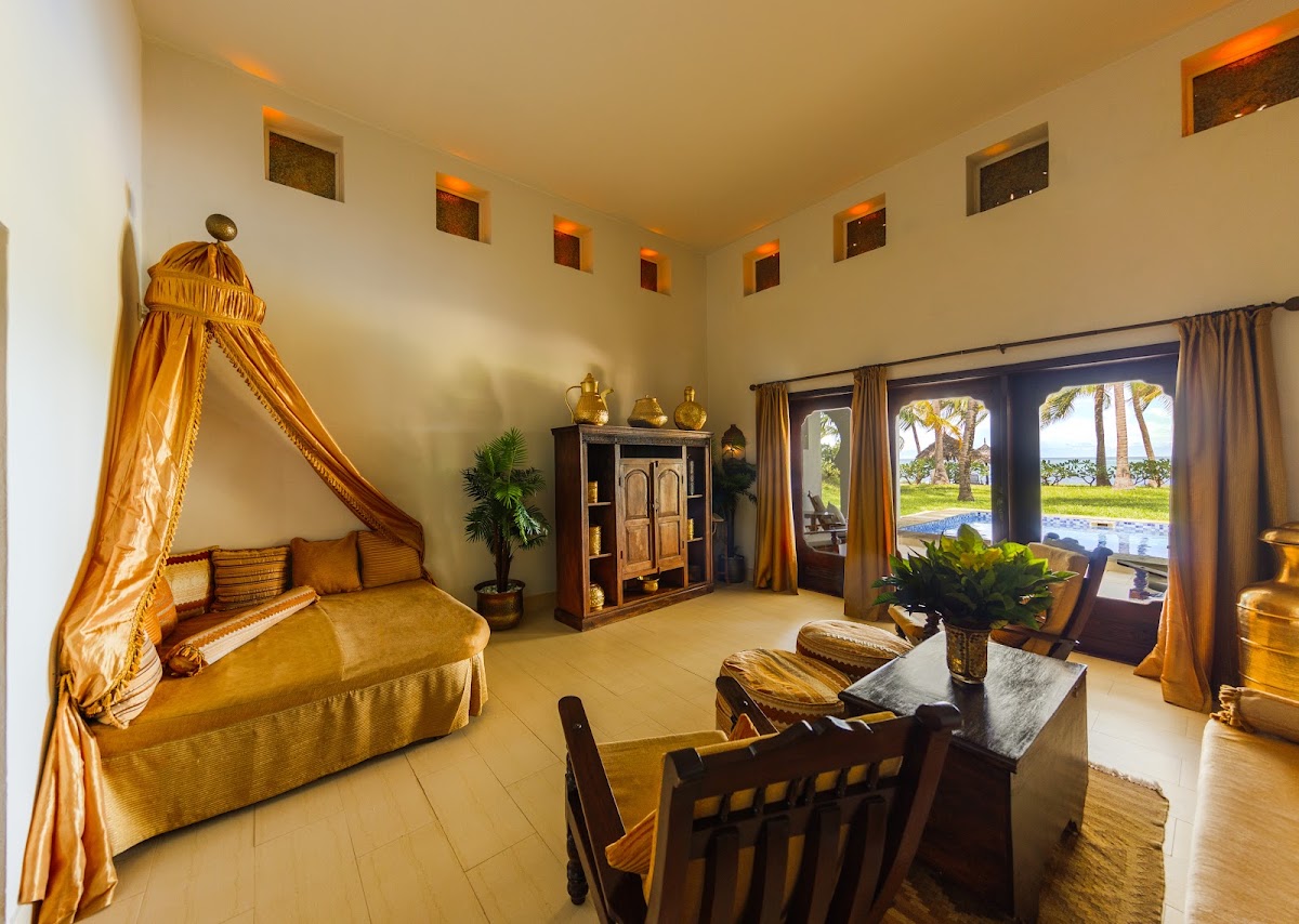 Luxury suite interior at Baraza Resort & Spa with gold canopied bed, wooden furnishings, and views of palm trees and lagoon through glass doors.