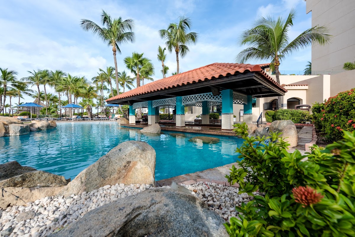 Turquoise pool with blue cabana structure, palm trees, and rocky landscaping at Barceló Aruba.