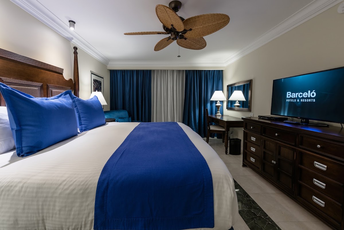 Barceló Aruba guest room with king bed dressed in blue and white linens, woven palm ceiling fan, dark wood furniture, and blue curtains flanking the windows.