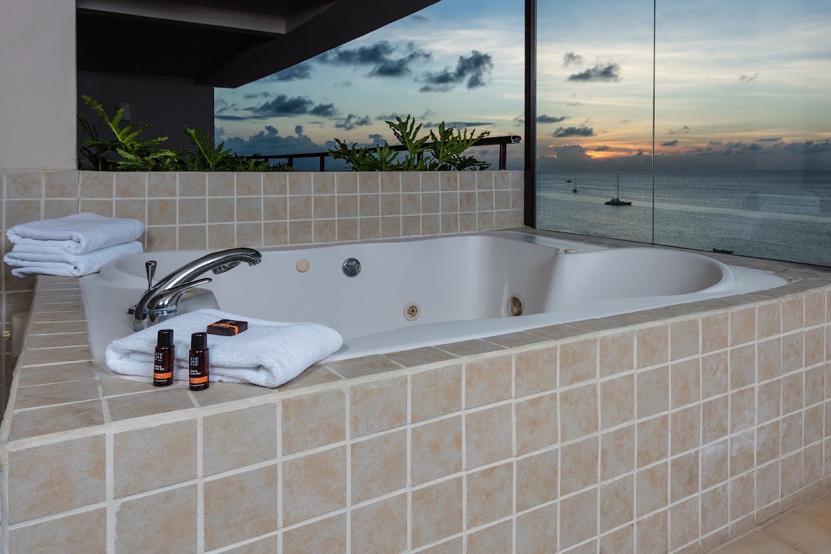 Bathroom with large soaking tub, white towels, and floor-to-ceiling windows overlooking a sailboat on the ocean at sunset.