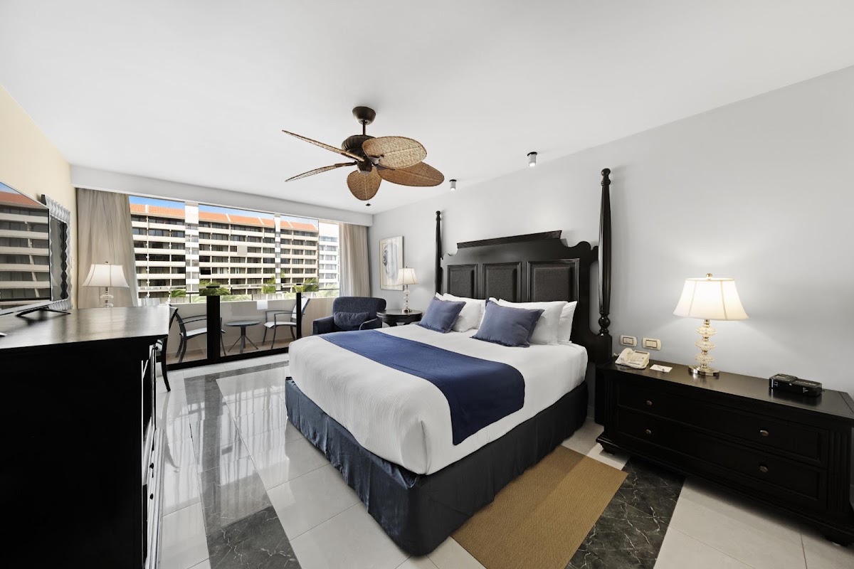 Resort bedroom with black four-poster bed, white linens with navy runner, ceiling fan, and windows overlooking high-rise buildings.