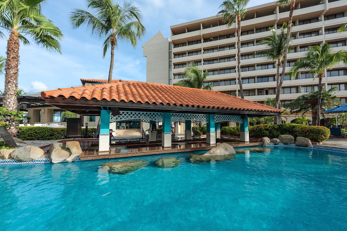 Turquoise pool with a teal and terra-cotta poolside cabana, palm trees, and multi-story resort building in the background.