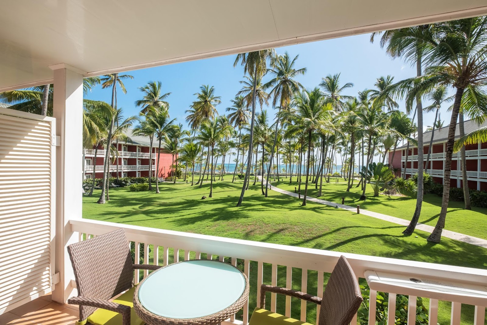 View from a beachfront balcony with wicker chairs and table overlooking manicured lawns, palm trees, and ocean at Barceló Bávaro Beach.