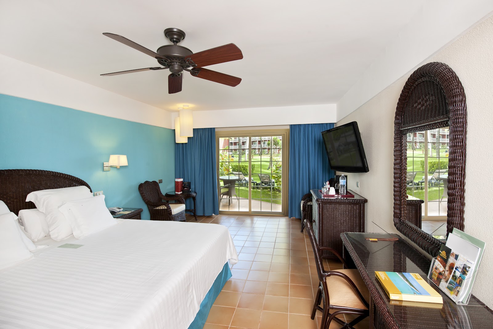Resort room with turquoise accent wall, wicker furniture, and glass doors opening to tropical grounds.