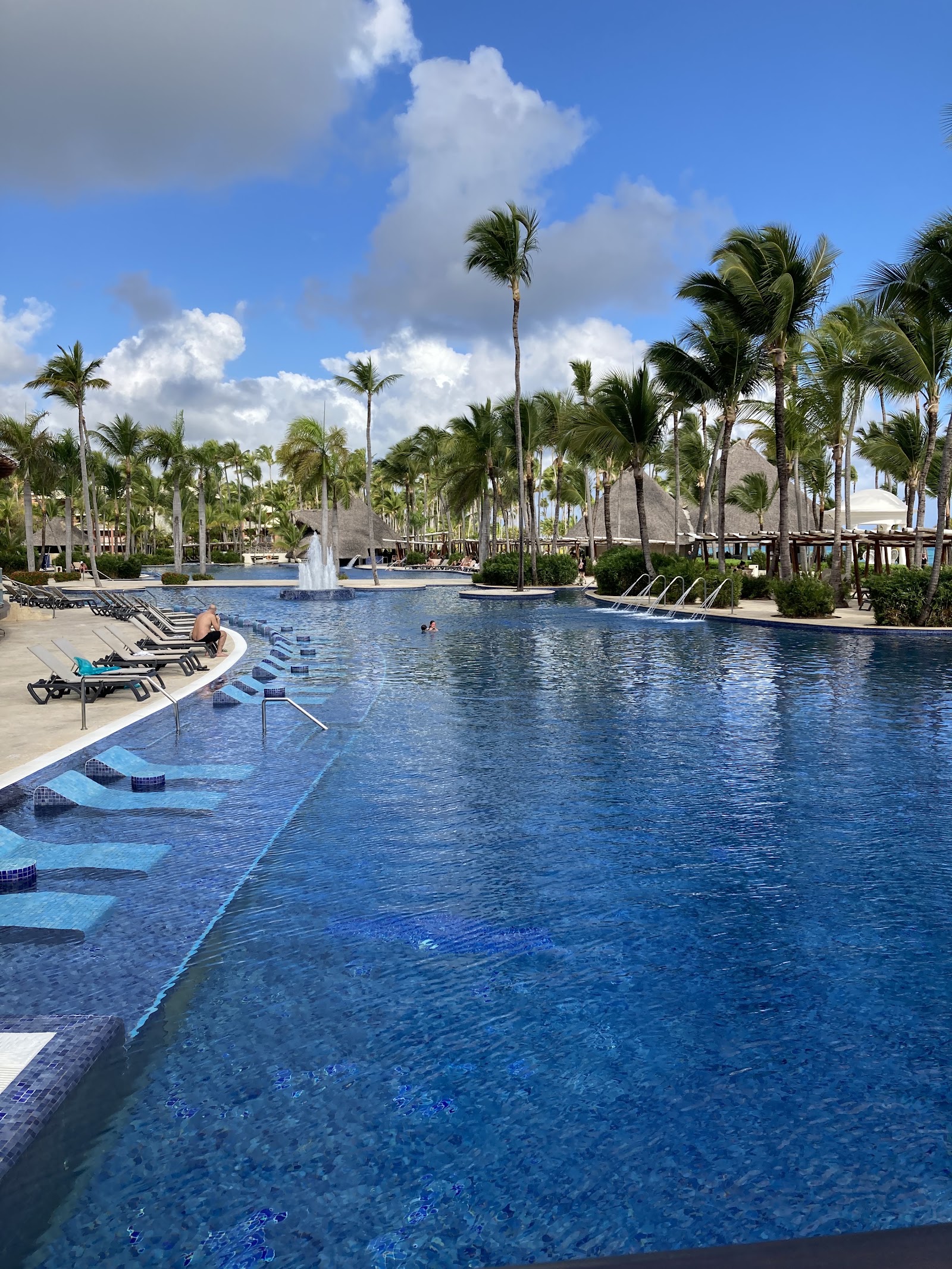 Large resort pool with fountains, lounge chairs along the edge, palm trees, and guests swimming under blue sky with white clouds.
