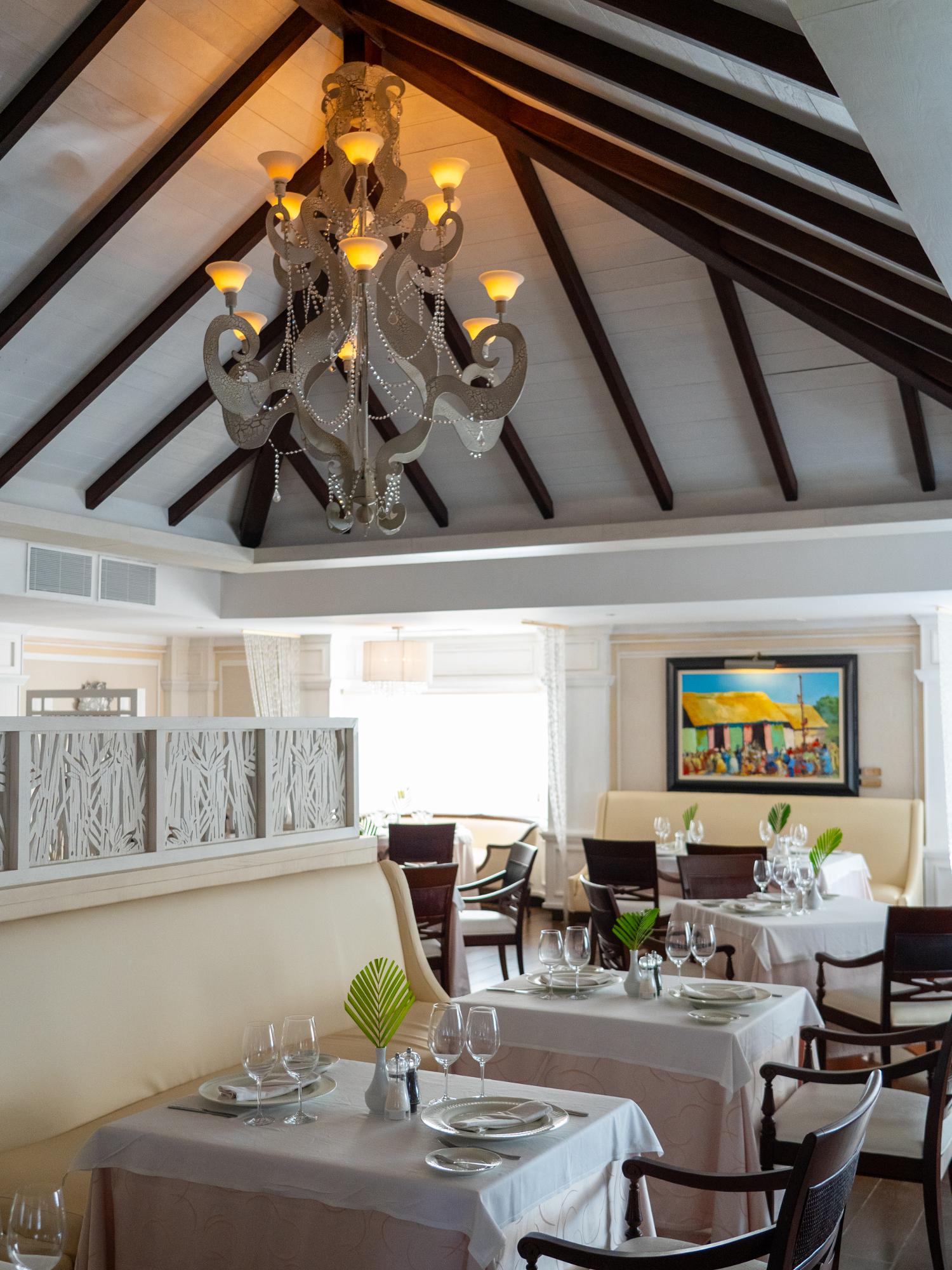 Upscale dining room with vaulted wood-beam ceiling, ornate chandelier, white-clothed tables set with glassware, and tropical artwork on cream-colored walls.