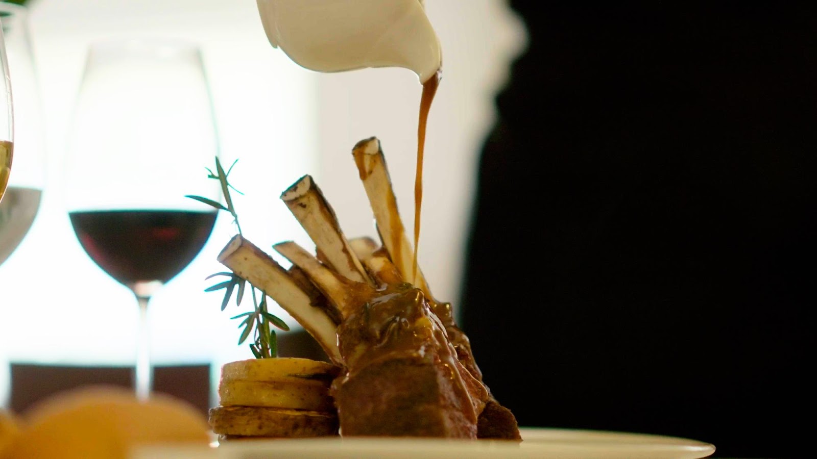 Sauce being drizzled over a plated lamb chop dish with rosemary garnish and potatoes, with wine glasses nearby.