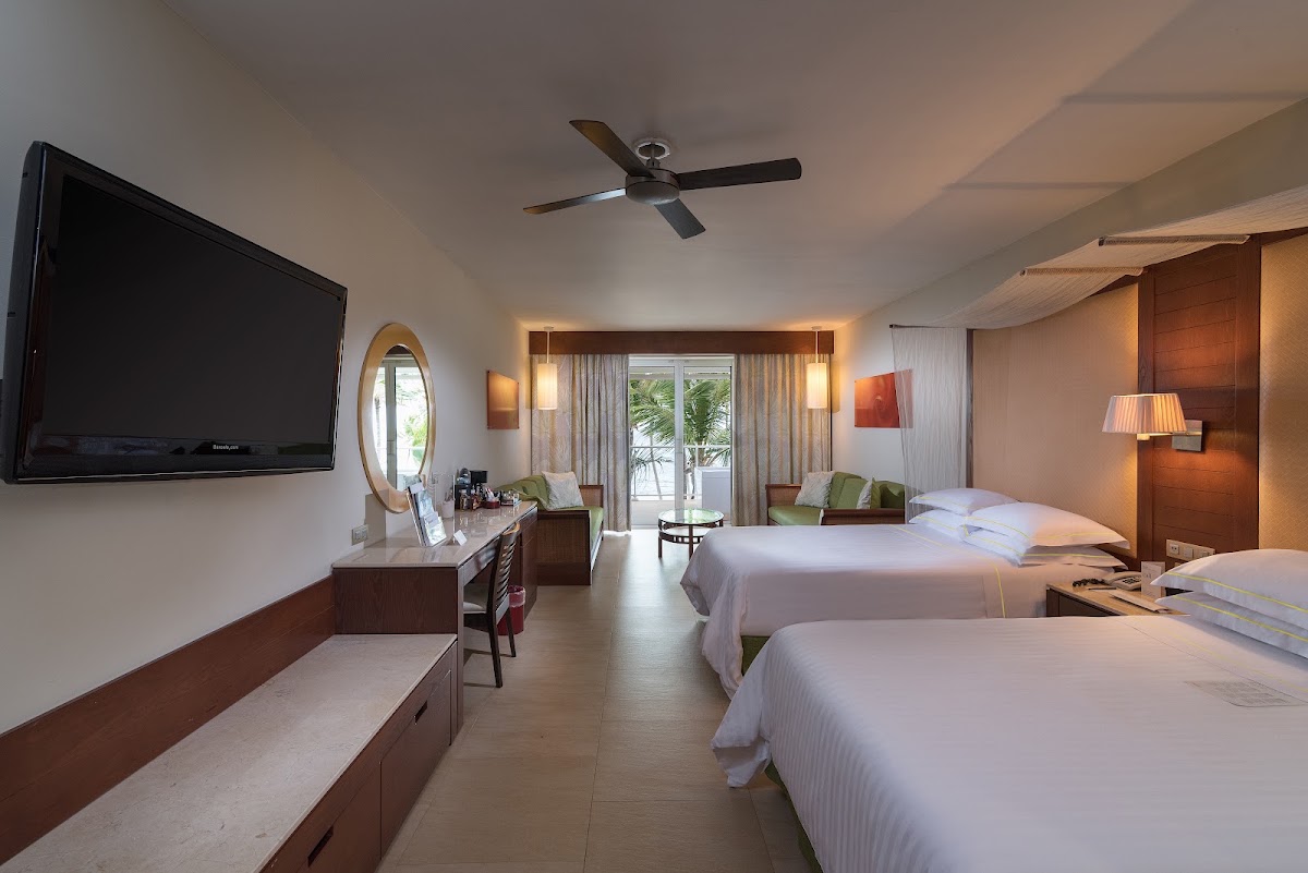 Resort guest room with two beds, wooden furnishings, flat-screen TV, and sliding doors opening to a terrace with tropical views.