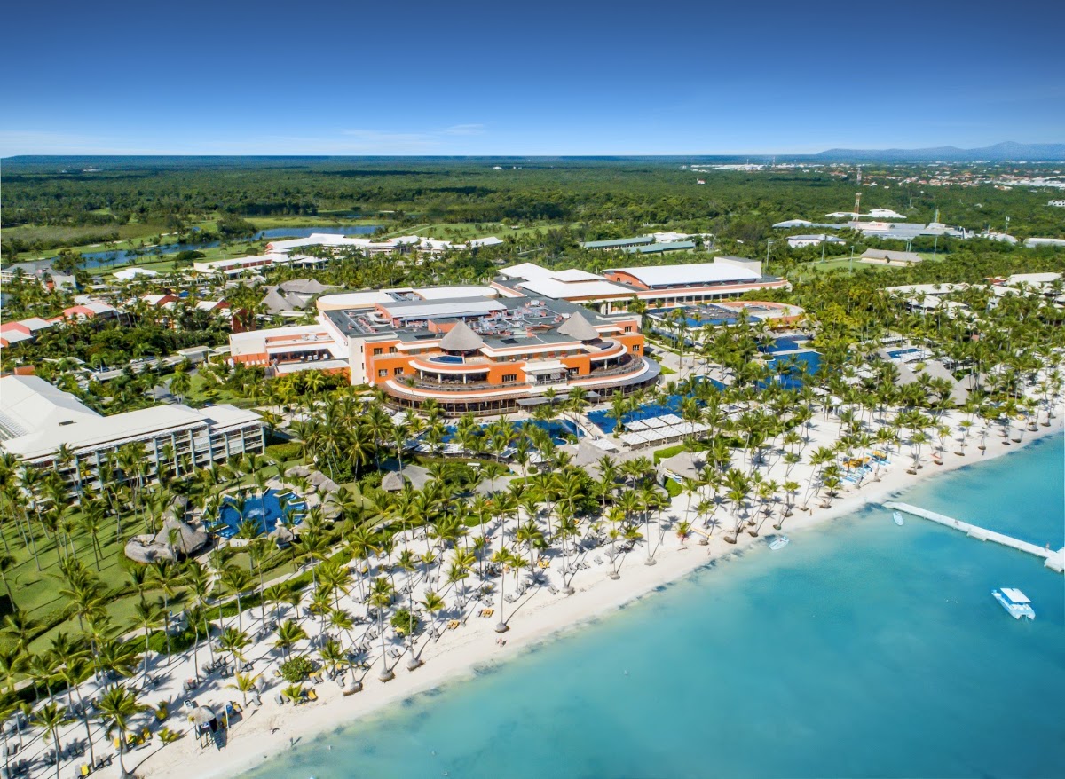Aerial view of Barceló Bávaro Palace beachfront resort with orange main building, palm-lined white sand beach, turquoise water, and lush green landscape beyond.