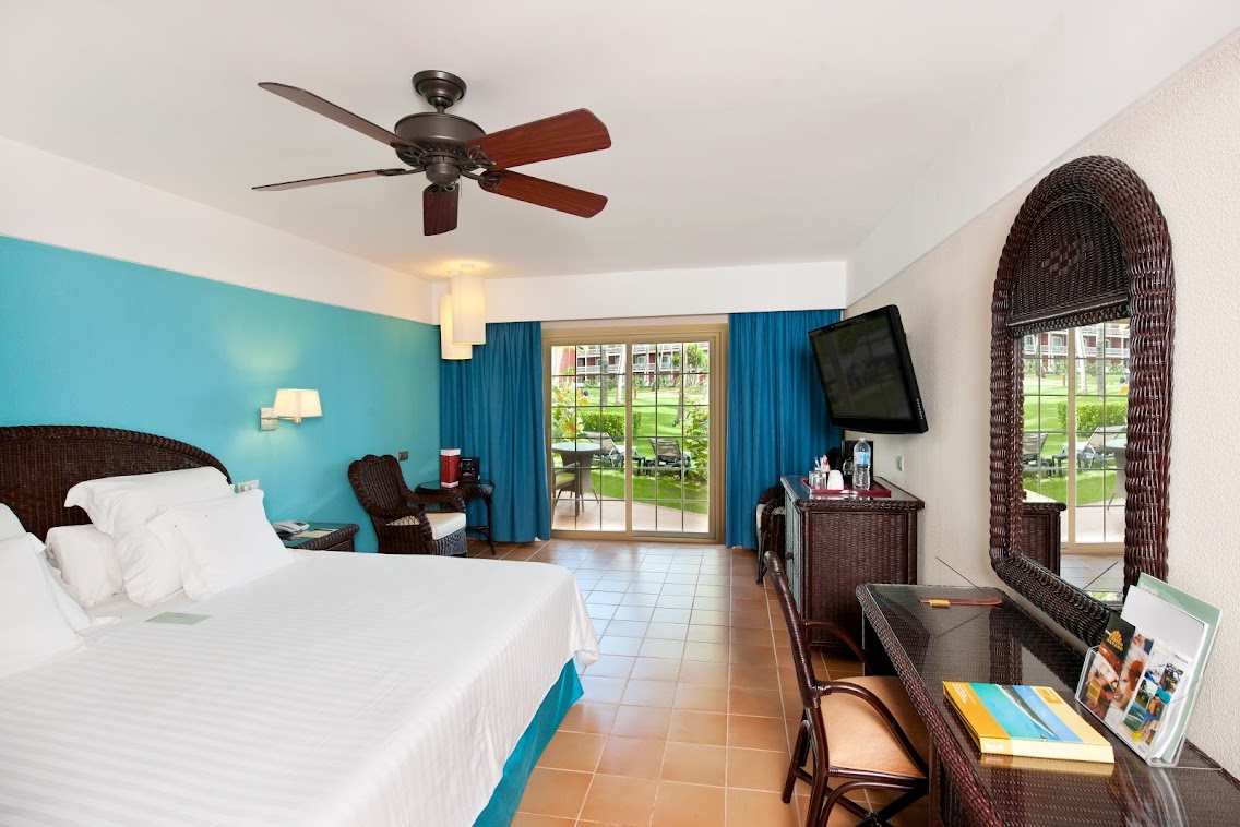 Resort guestroom with turquoise accent wall, wicker furniture, and glass doors opening to tropical grounds.