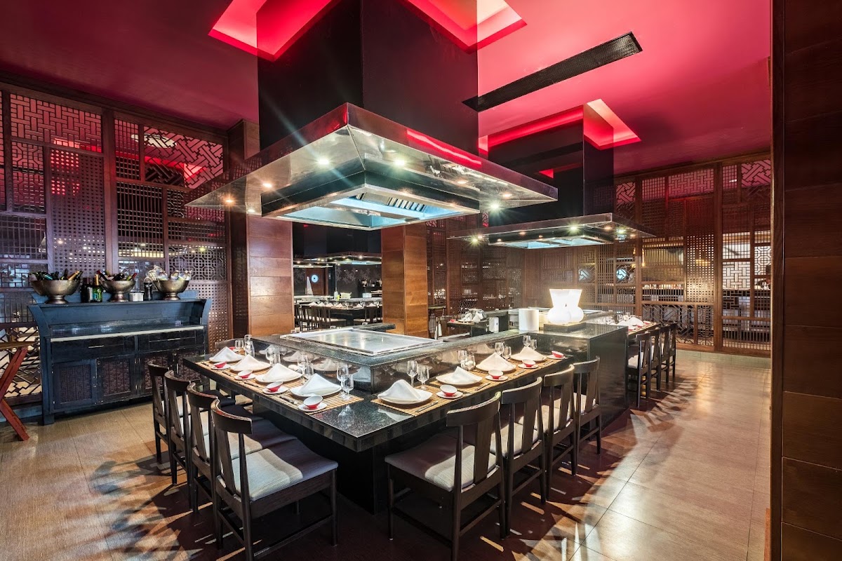 Teppanyaki restaurant with pink neon ceiling, open kitchen with steel hood, black dining tables set with place settings, and decorative lattice screens.