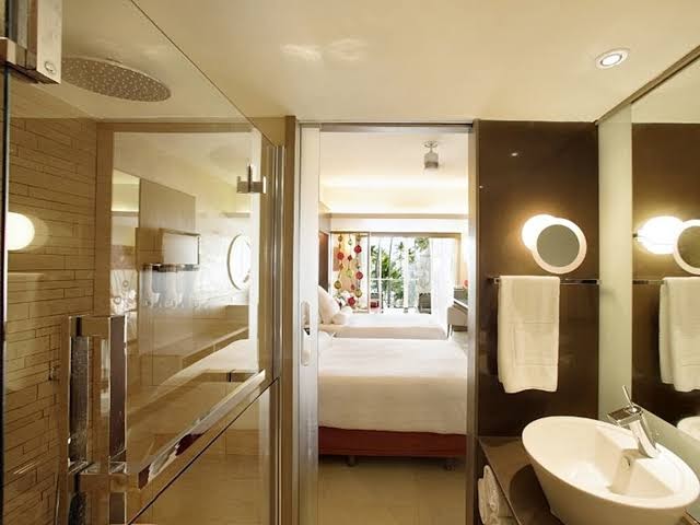 Bathroom with glass shower enclosure, round illuminated mirrors, and white sink, opening to bedroom with bed and garden view beyond.