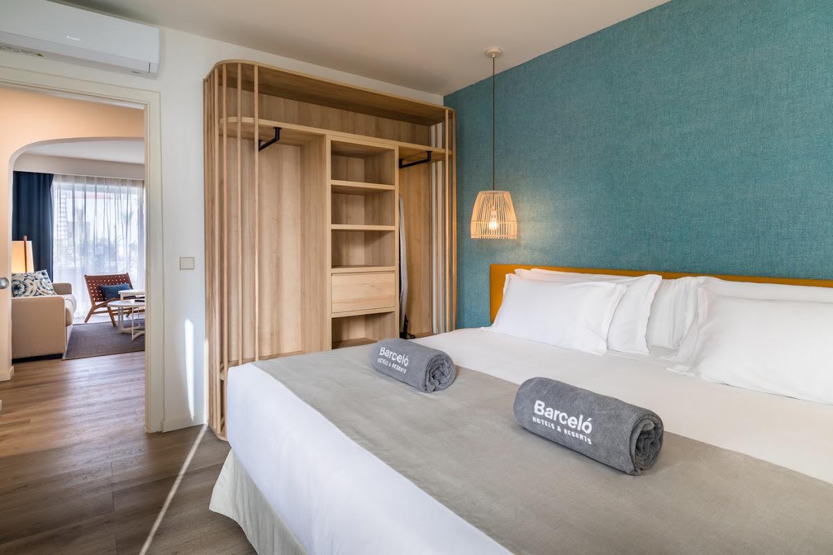 Barceló Fuerteventura Royal Level bedroom with queen bed, teal accent wall, wooden wardrobe, and open doorway to living area.