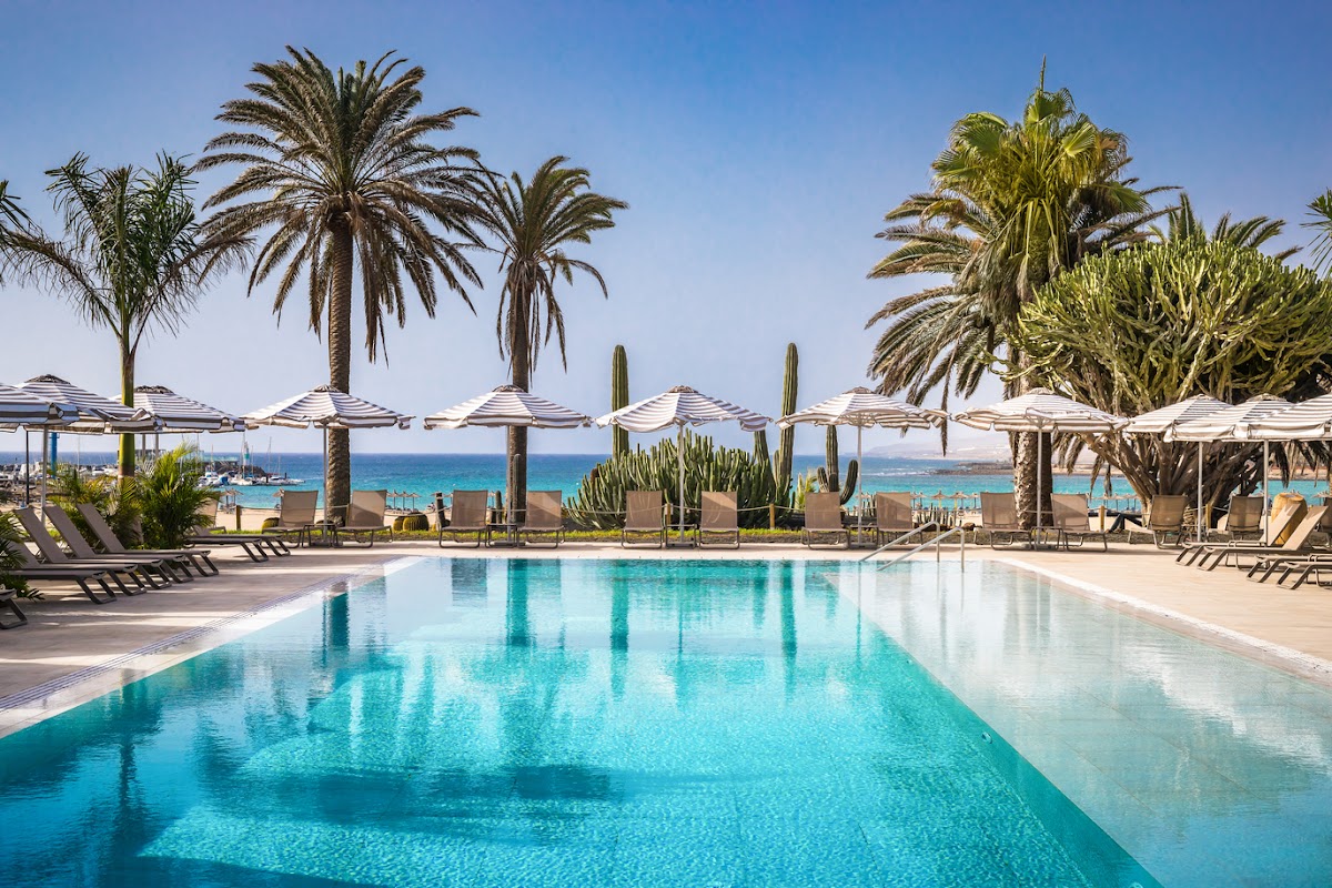 Resort pool lined with white umbrellas and lounge chairs, palm trees, and turquoise ocean beyond under clear sky.