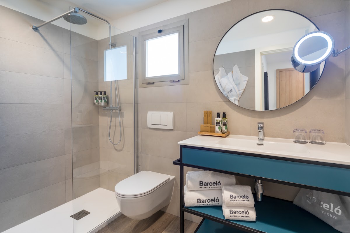 Barceló Fuerteventura Royal Level bathroom with walk-in shower, round mirror, white sink vanity with teal accents, and branded toiletries.