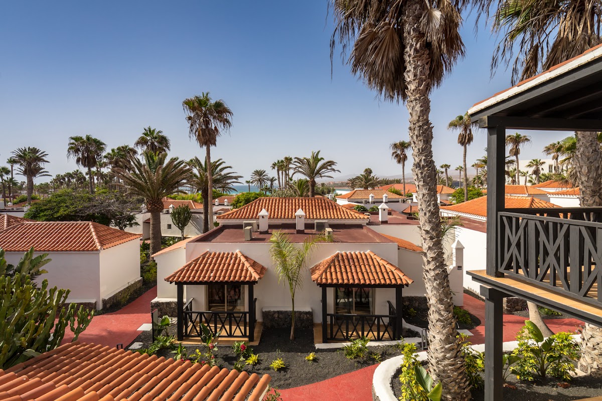 Aerial view of Barceló Fuerteventura Royal Level showing white buildings with terracotta roofs, palm trees, and ocean in the distance under clear blue sky.