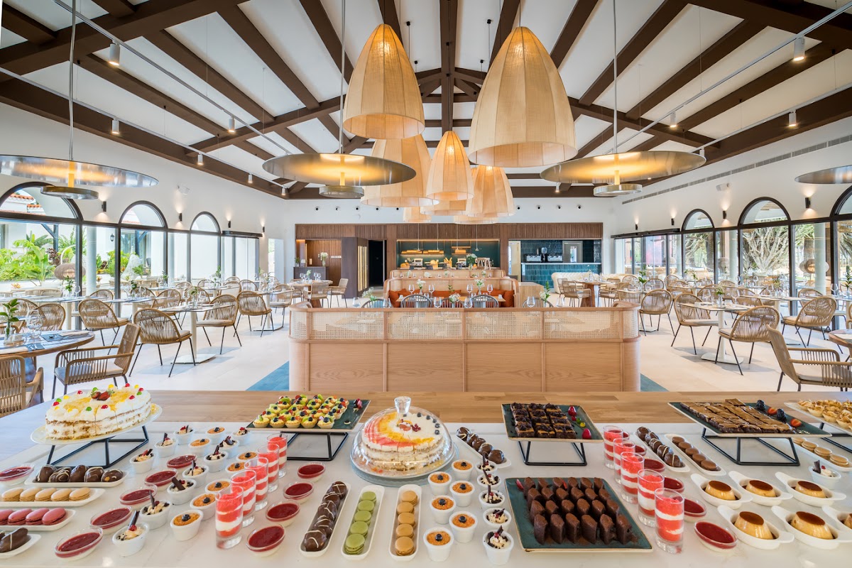 Barceló Fuerteventura Royal Level dining hall with dessert buffet in foreground, wooden pendant lights, beam ceiling, and woven chairs arranged throughout the bright space.