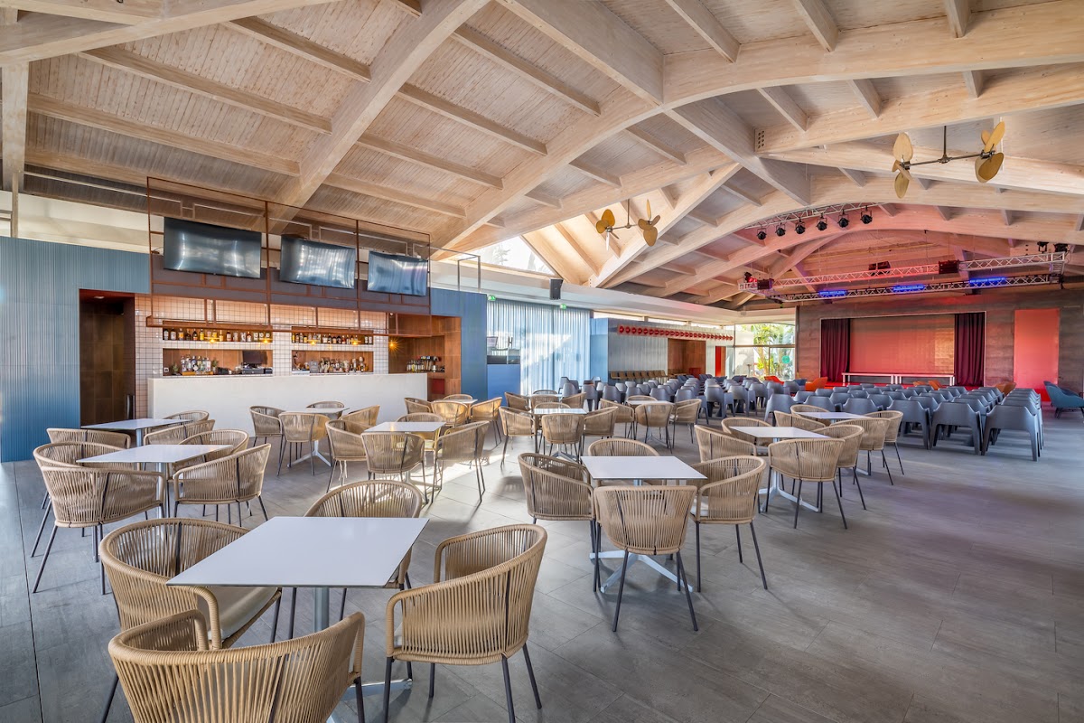 Open-air dining pavilion with woven chairs around white tables, exposed wooden beam ceiling, bar counter, and stage area with red backdrop at Barceló Fuerteventura Royal Level.