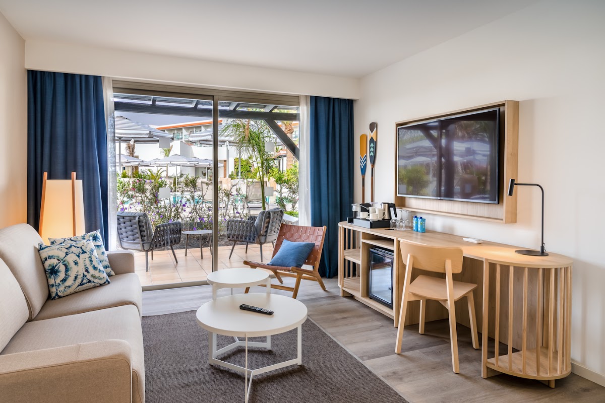 Modern suite with beige seating, wooden desk, and glass doors opening to a furnished terrace with tropical plants at Barceló Fuerteventura Royal Level.