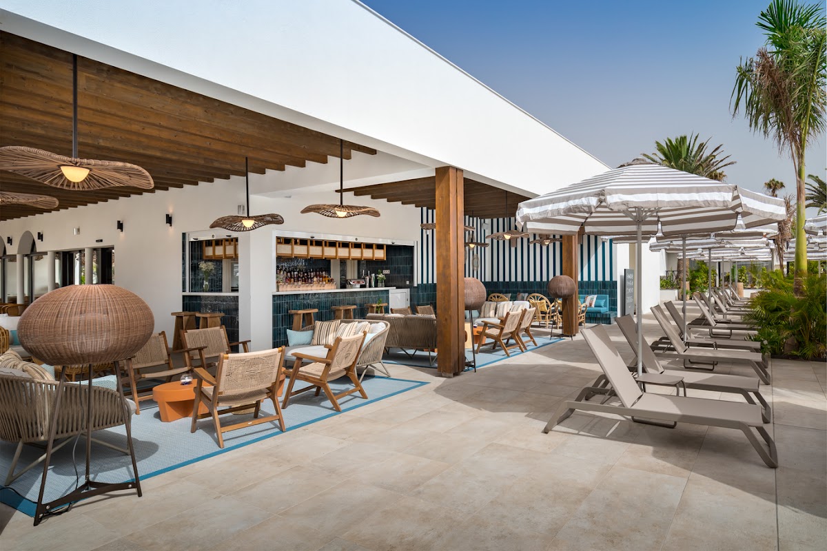 Modern poolside bar and lounge area at Barceló Fuerteventura Royal Level with woven seating, striped umbrellas, and palm trees under clear sky.