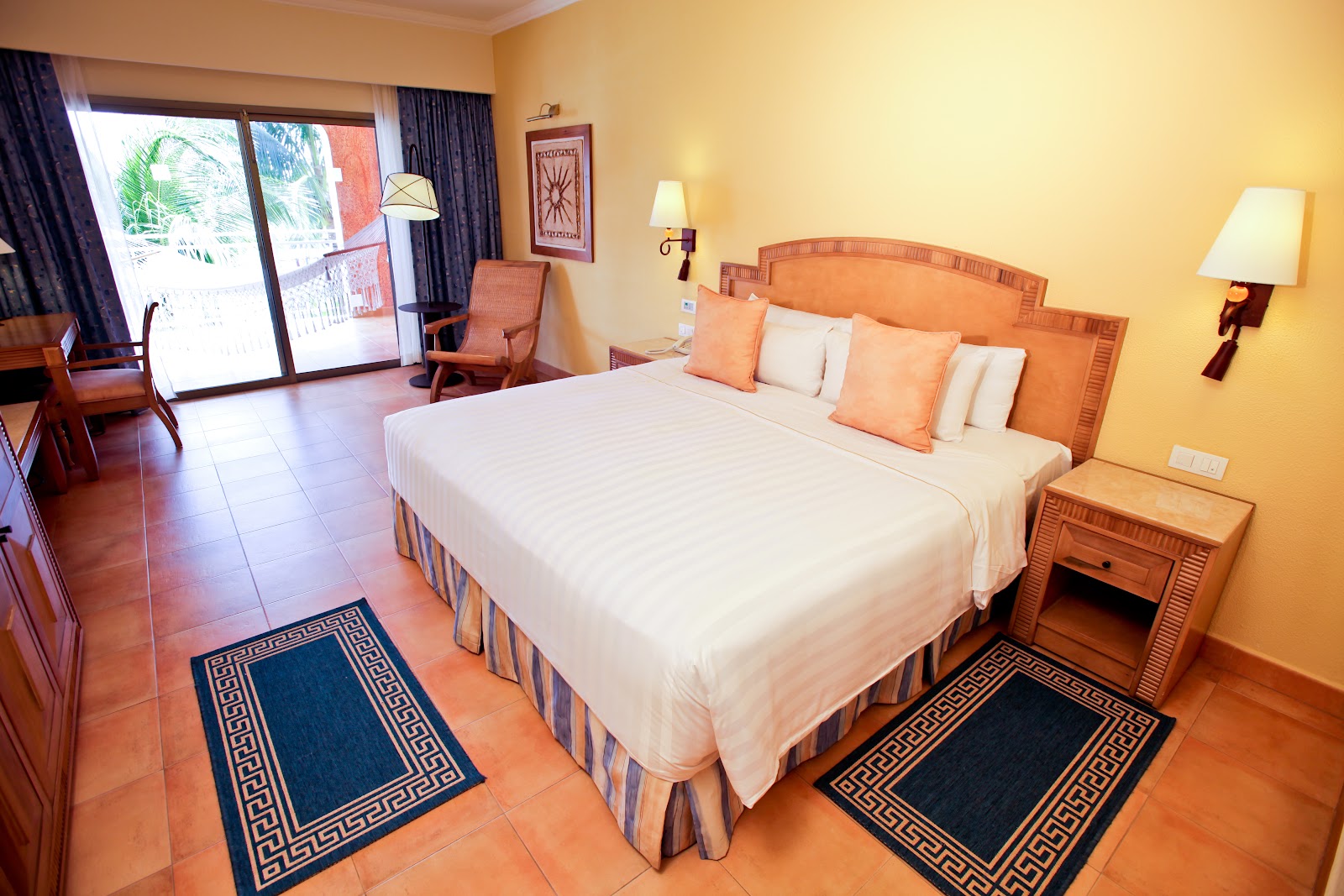 Resort bedroom with queen bed, orange headboard, cream bedding, terra cotta tile floor, yellow walls, and sliding glass doors opening to a balcony.