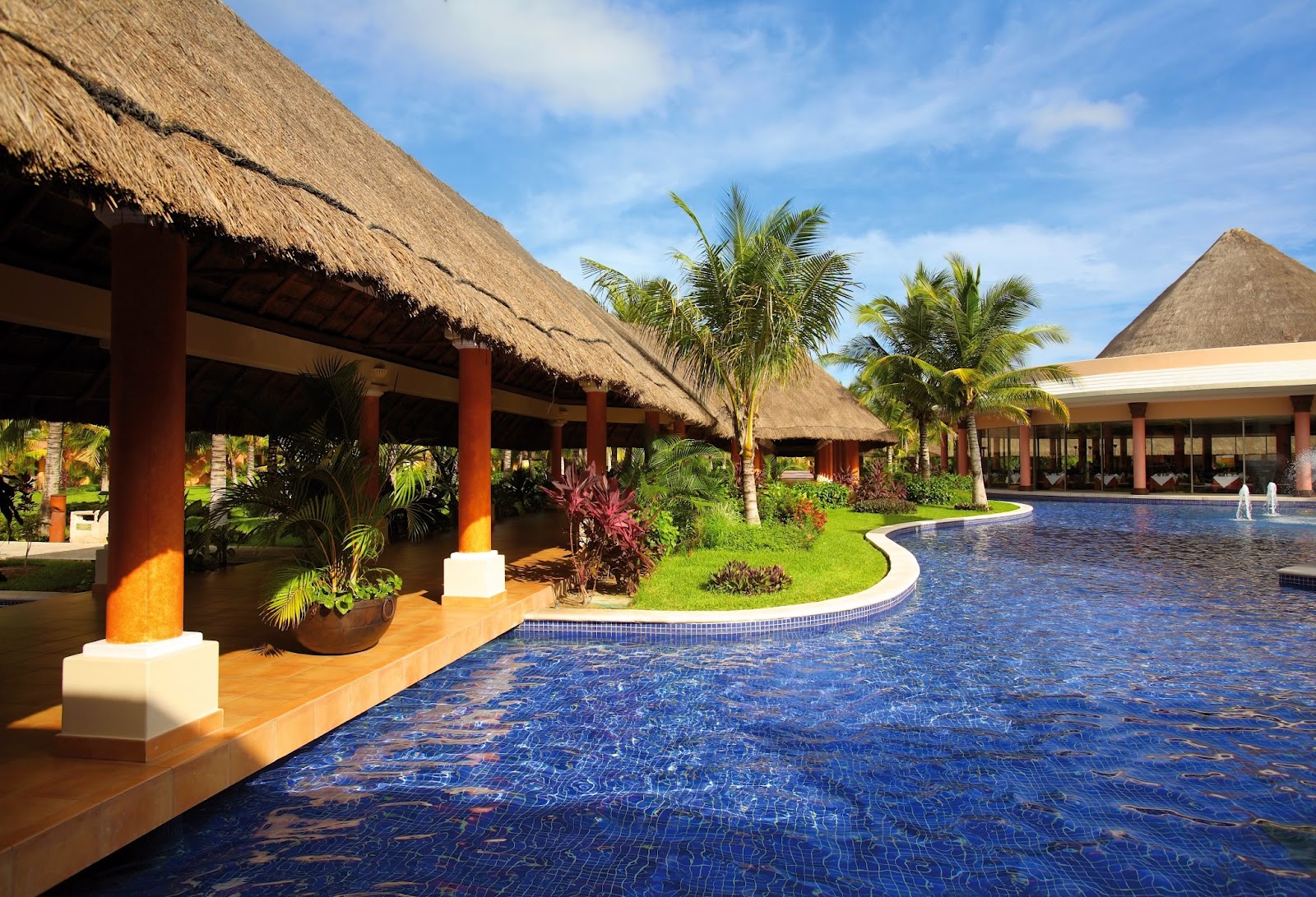Thatched-roof buildings with red columns frame a curved pool surrounded by palm trees and tropical landscaping under blue sky.