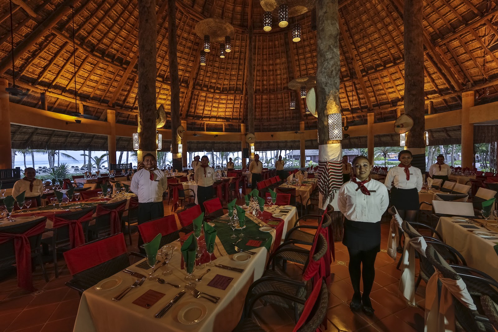 Thatched-roof dining hall with wooden beams at Barceló Maya Colonial, staff in formal attire standing among tables set with white linens and red chairs, ocean view through open sides.