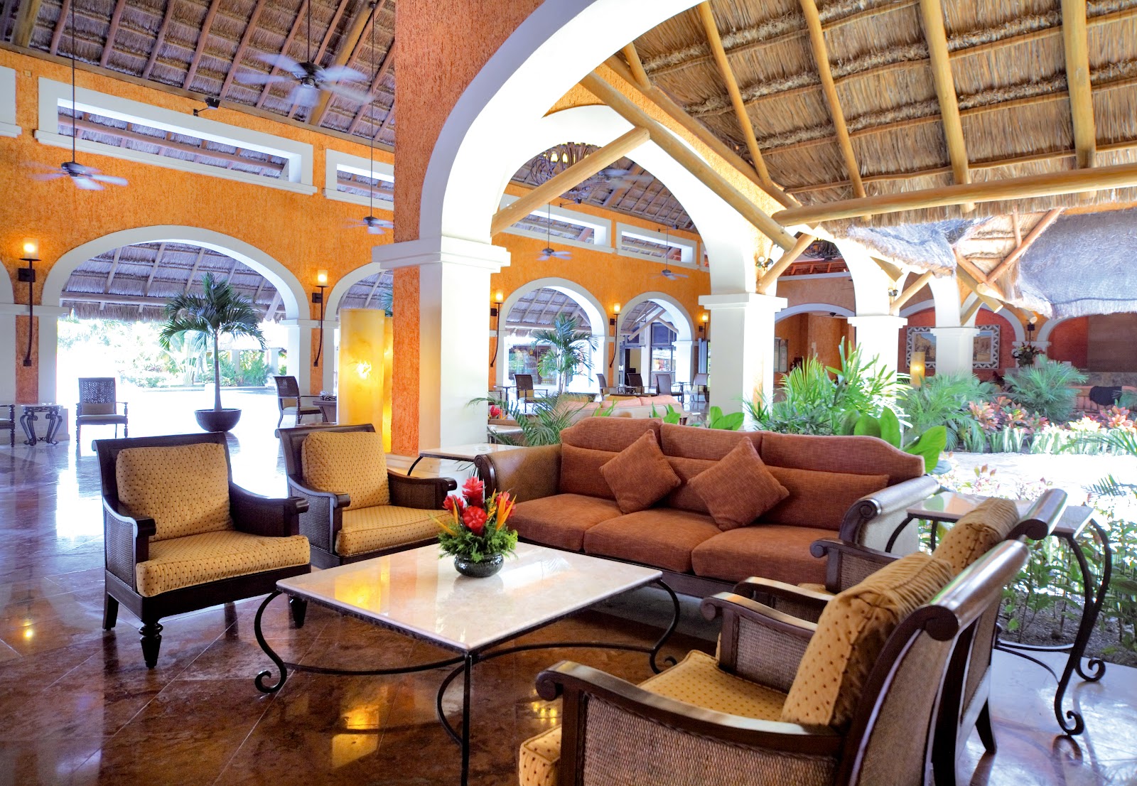 Orange-walled resort lobby with arched doorways, wooden beam ceiling, brown upholstered seating, potted palms, and natural light streaming through openings.
