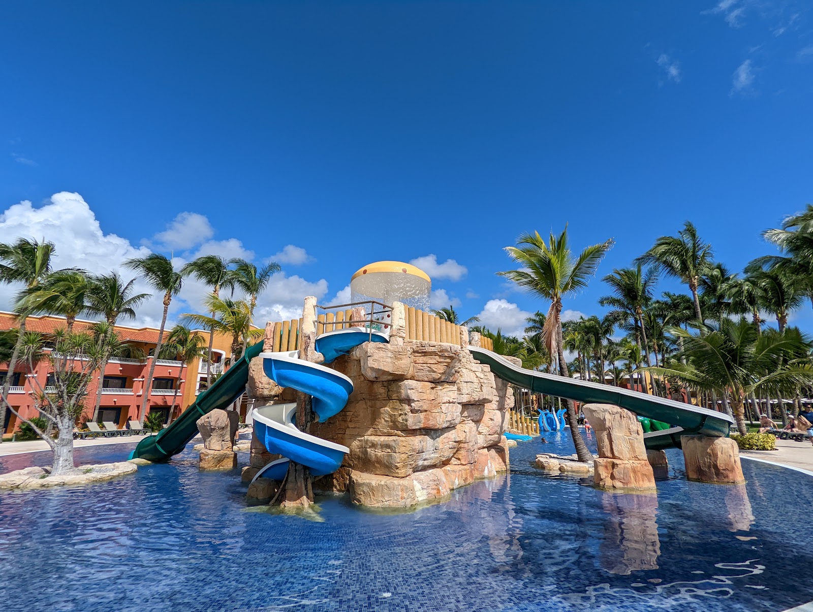 Water slide tower with blue slides spiraling down rocky structure in resort pool area surrounded by palm trees and colorful buildings.