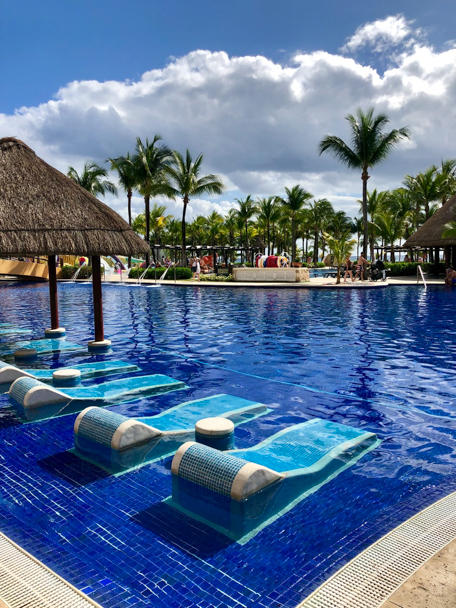 Floating exercise platforms in a resort pool with palm trees and thatched-roof structures along the beach at Barceló Maya Grand Resort.
