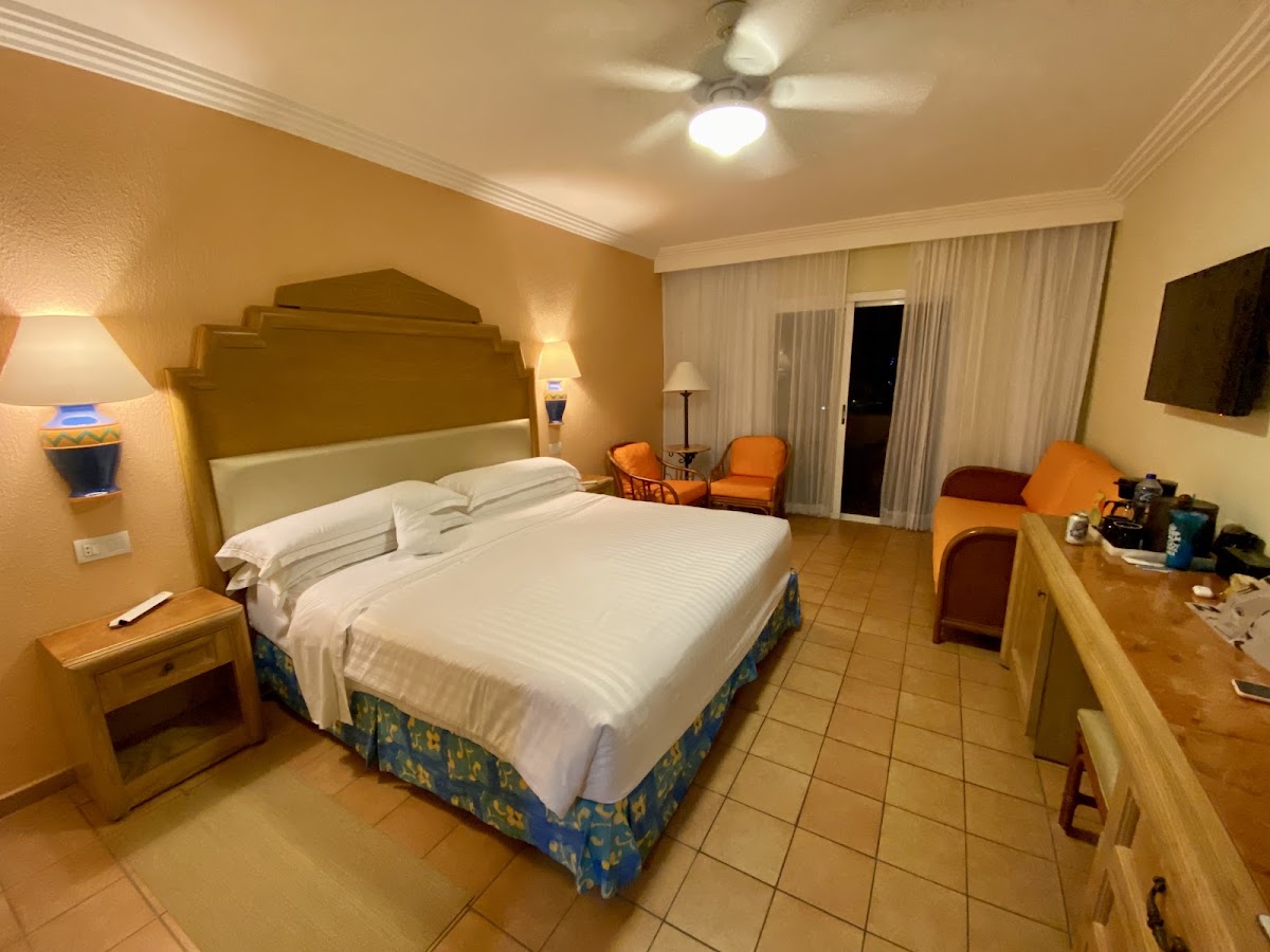 Resort bedroom with queen bed, gold headboard, orange accent chairs, and tile flooring, lit by ceiling fan and lamps.