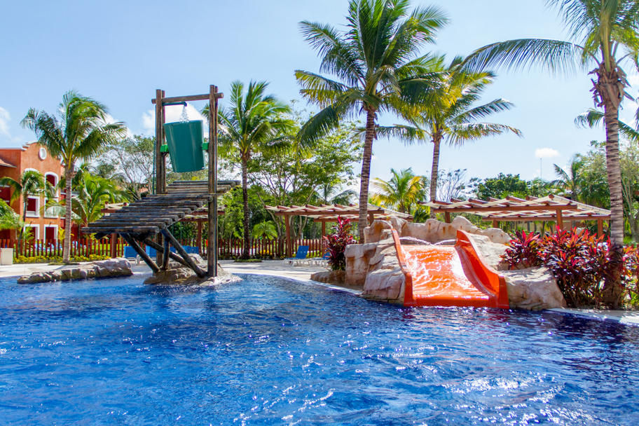 Water slides and play structures in a bright blue pool surrounded by palm trees and red umbrellas at Barceló Maya Grand Resort.