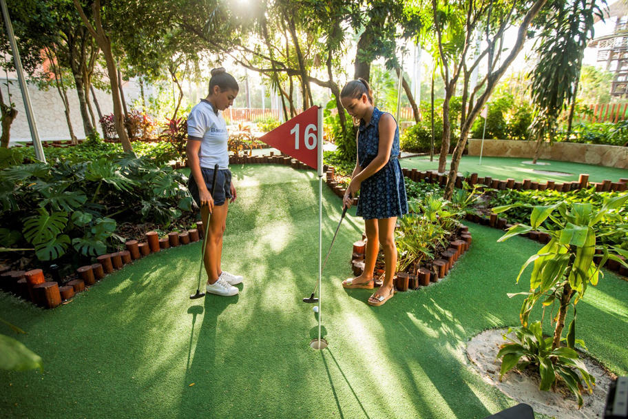Two children playing mini golf on a shaded green course lined with tropical plants and a red flag marking hole 16.