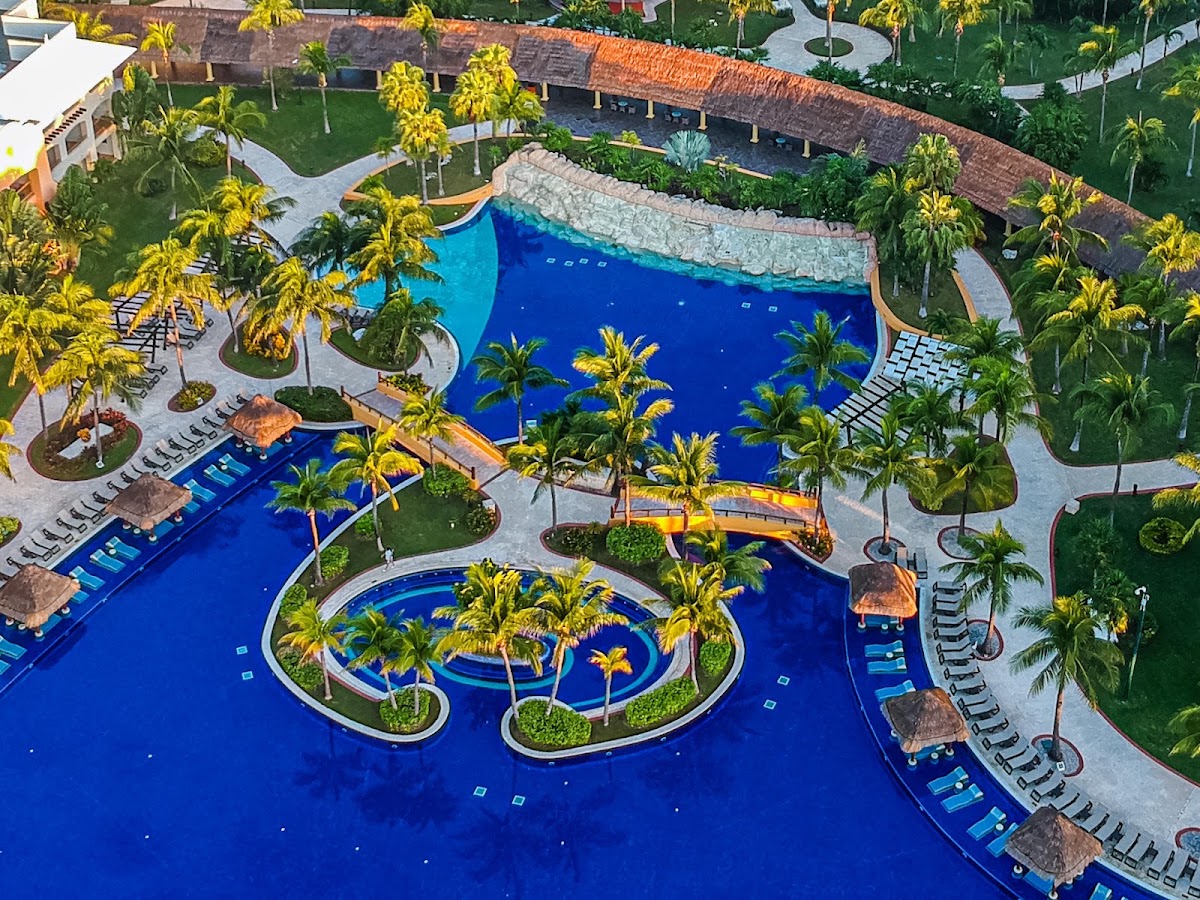 Aerial view of Barceló Maya Grand Resort's pool complex with multiple blue pools, palm trees, lounge chairs, and thatched-roof cabanas surrounded by manicured lawns.