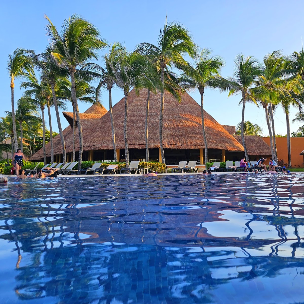 Resort pool with thatched-roof palapa structure, palm trees, and lounge chairs reflected in still blue water.