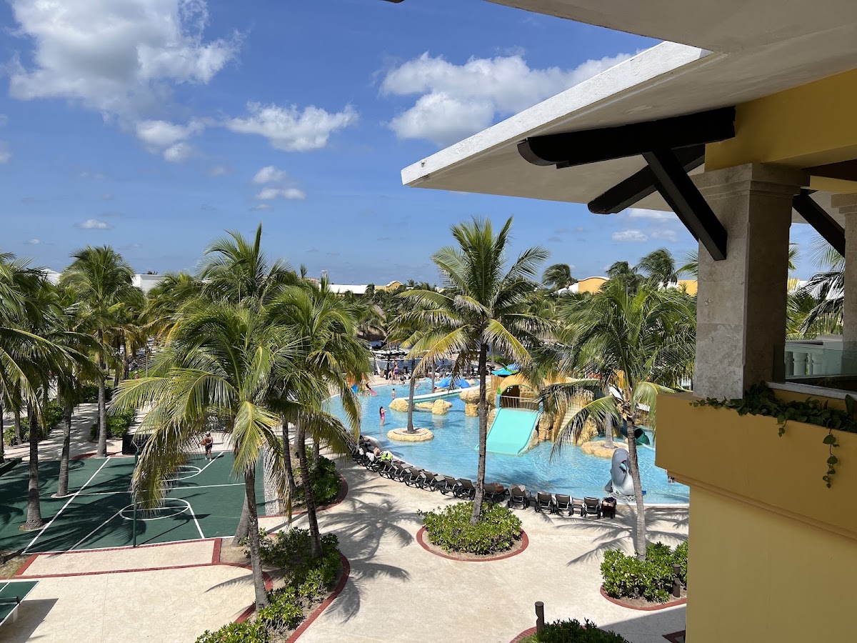 View from a shaded terrace over palm trees and a turquoise pool with rock features at Barceló Maya Grand Resort.
