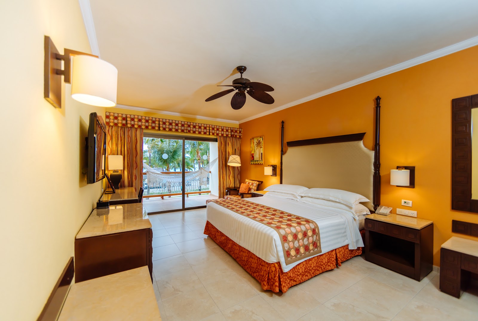 Resort bedroom with orange walls, four-poster bed with patterned runner, dark wood furniture, and sliding glass doors opening to ocean view.