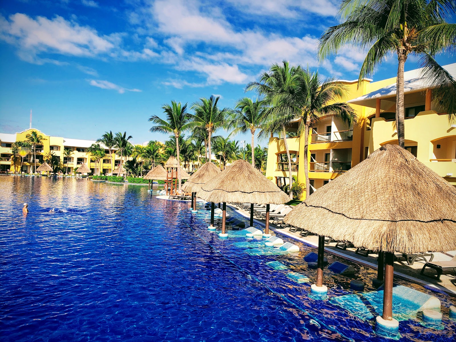 Yellow resort buildings with thatched umbrellas line a lagoon pool surrounded by palm trees under blue sky.